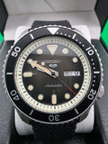 A Seiko Automatic dive watch with a black dial, black bezel, and black textured strap, displayed inside its white presentation box.