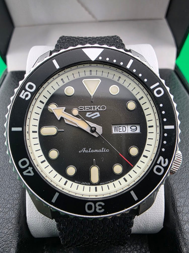A Seiko Automatic dive watch with a black dial, black bezel, and black textured strap, displayed inside its white presentation box.